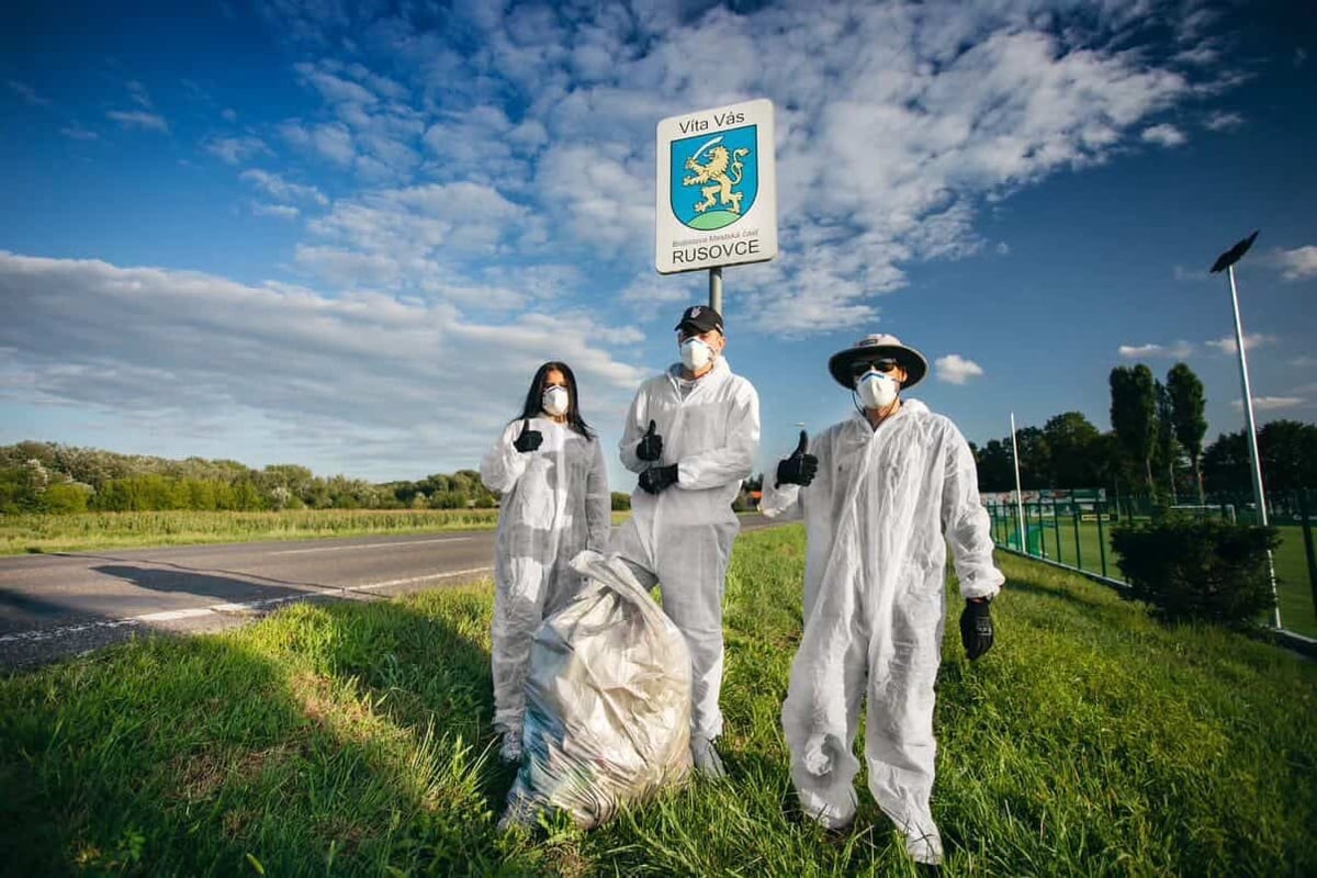 Three people in white protective suits holding a sign with a coat of arms on a grassy field under blue sky
