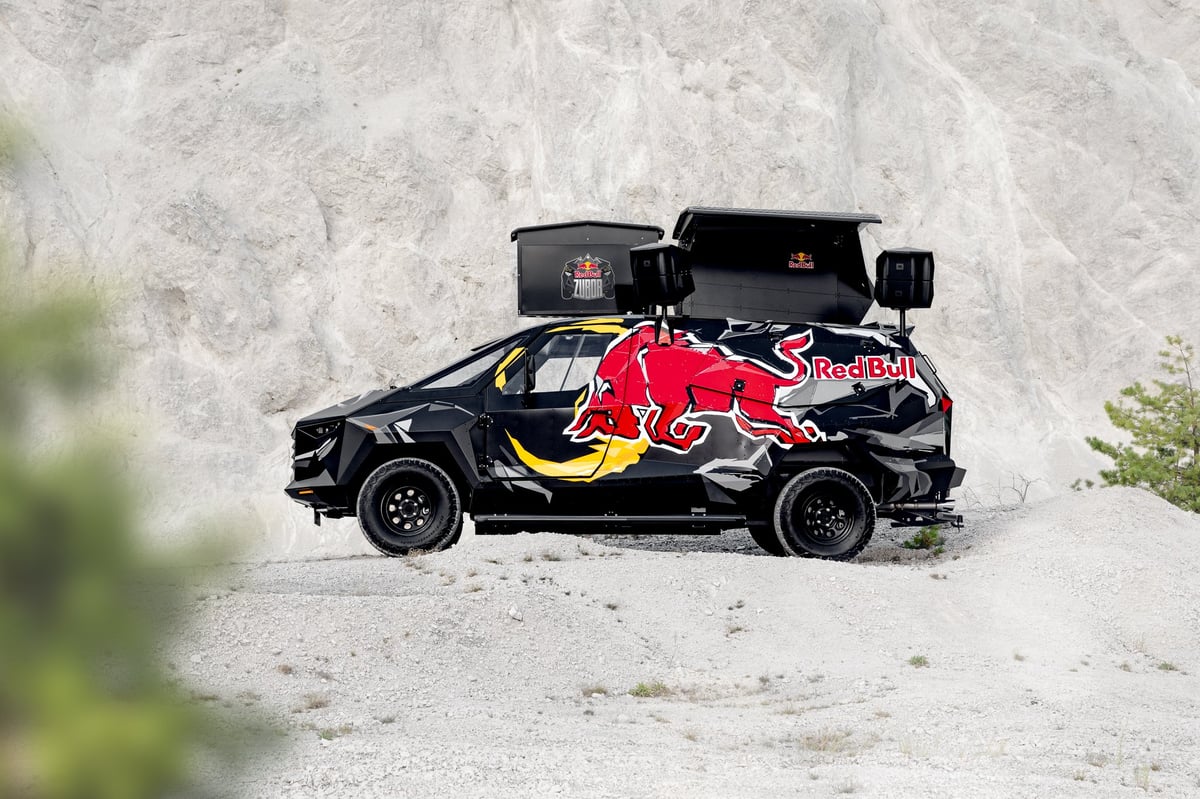 Black rally car with Red Bull graphics and equipment mounted on roof, parked against white stone wall