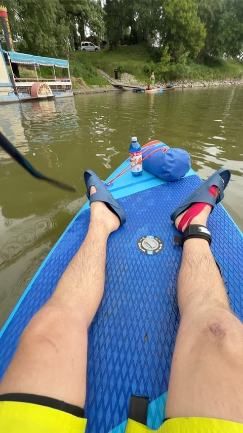 First-person view of paddler on blue stand-up paddleboard on a calm river with trees and docks in the background