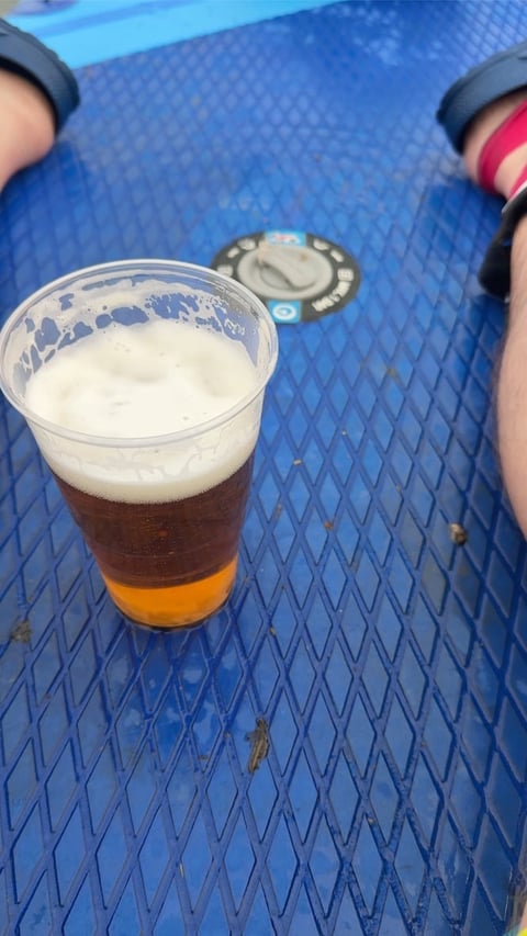 Person holding a plastic cup of dark beer with foam head on a blue textured surface next to a bottle cap