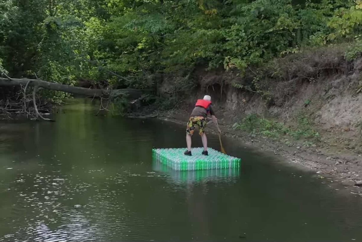 Person in red helmet standing on a green and blue raft floating on a calm river surrounded by green forest and trees