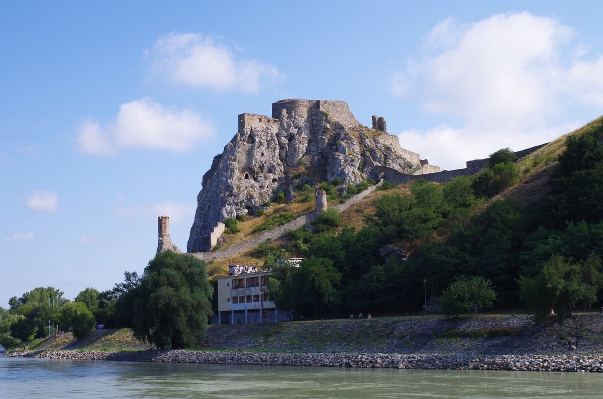 Ancient castle ruins perched on rocky hilltop overlooking a river with houses nestled on the green slope below