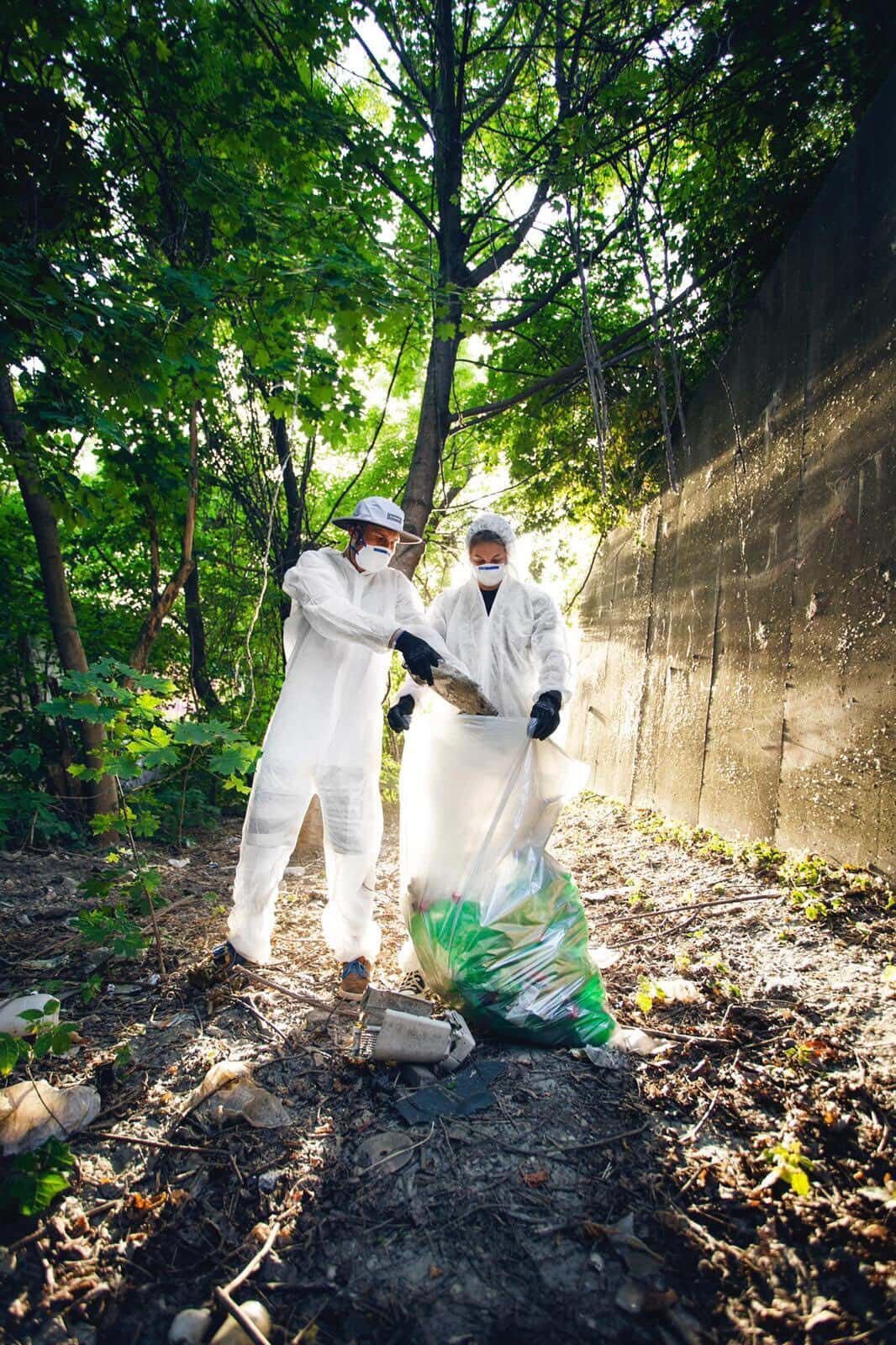 Two workers in white protective suits and masks collecting waste into a green plastic bag in a shaded outdoor alley